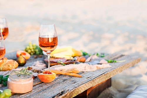 Ein elegantes Picknick am Strand mit einem Glas Roséwein, frischen Früchten und feinen Delikatessen auf einem rustikalen Holztisch.